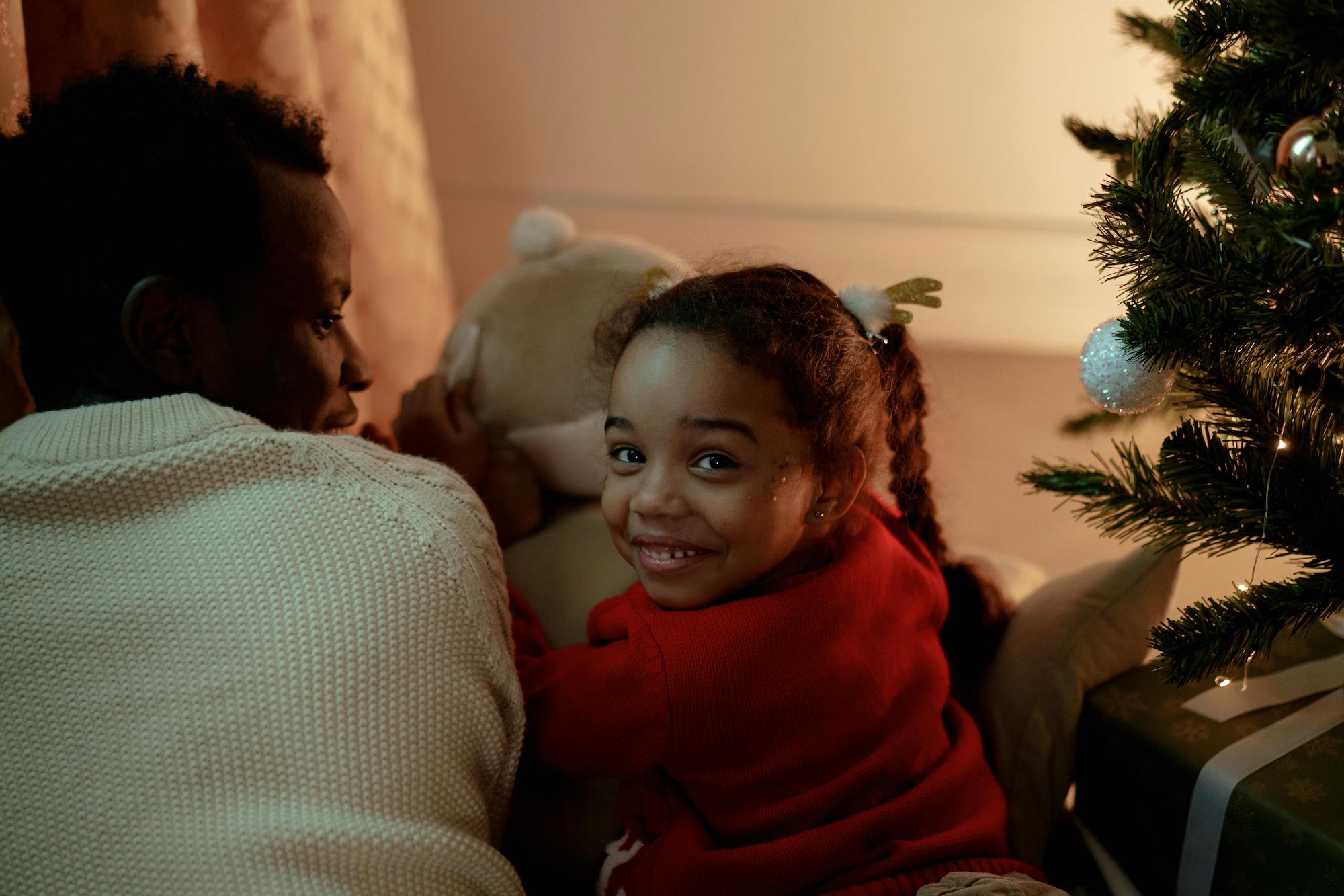 Little girl in red shirt next to Christmas tree with her dad