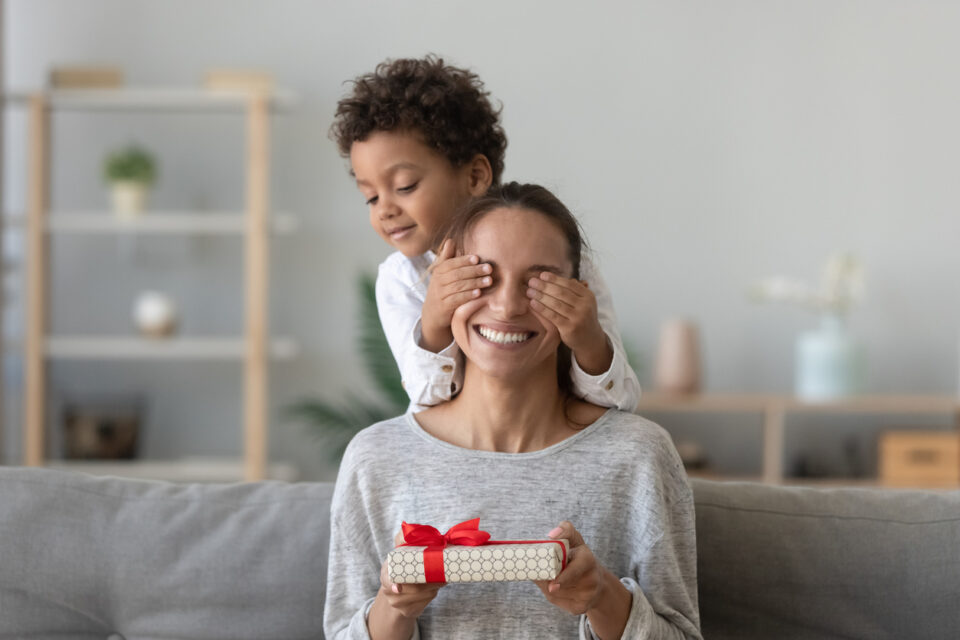 Happy excited woman sitting on couch in living room with covered by playful small preschool kid boy hands eyes, holding wrapped gift box, feeling curious. Birthday or Mothers day celebration concept.