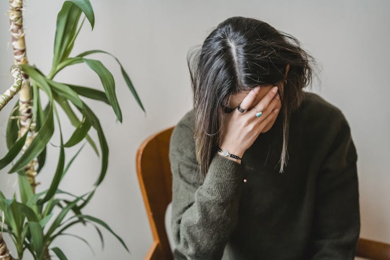 Tired mom in stylish sweater covering face with hand while siting on chair at home