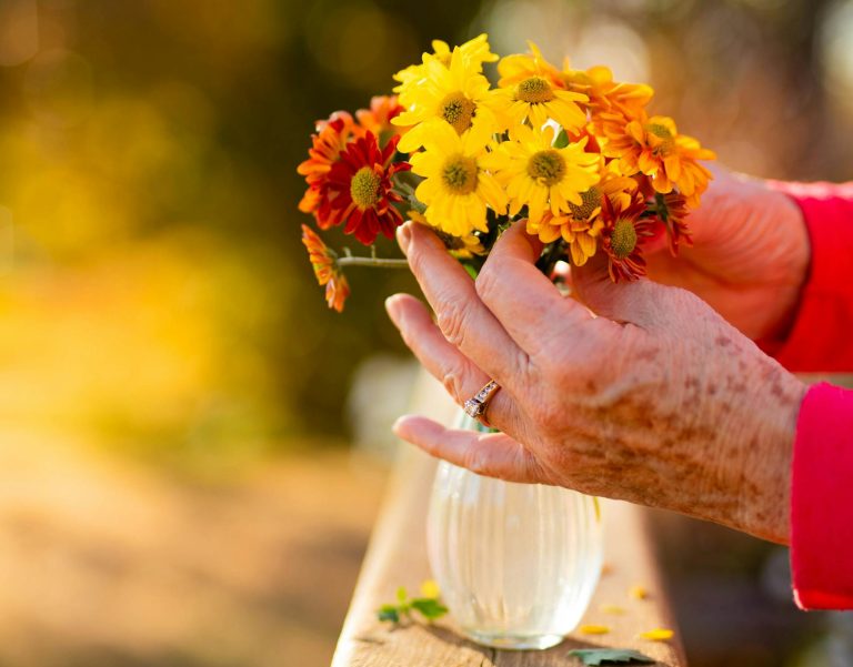 Fun Things about getting older - Senior hands arranging yellow and orange flowers in a vase with a blurred outdoor background.